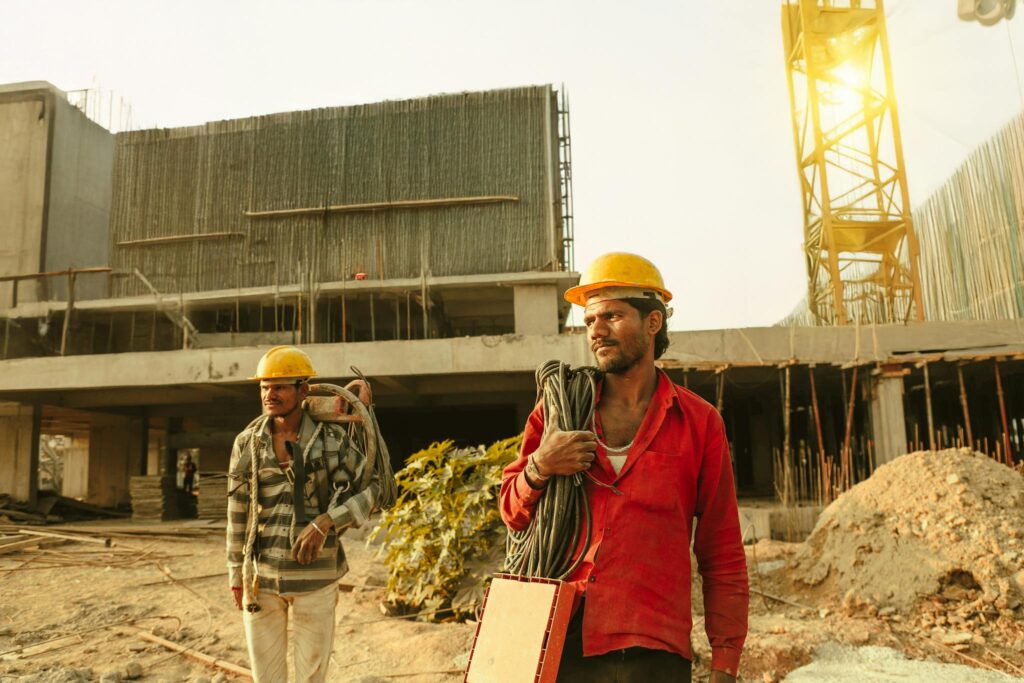 Two construction workers wearing helmets at a site in India with a crane and building.